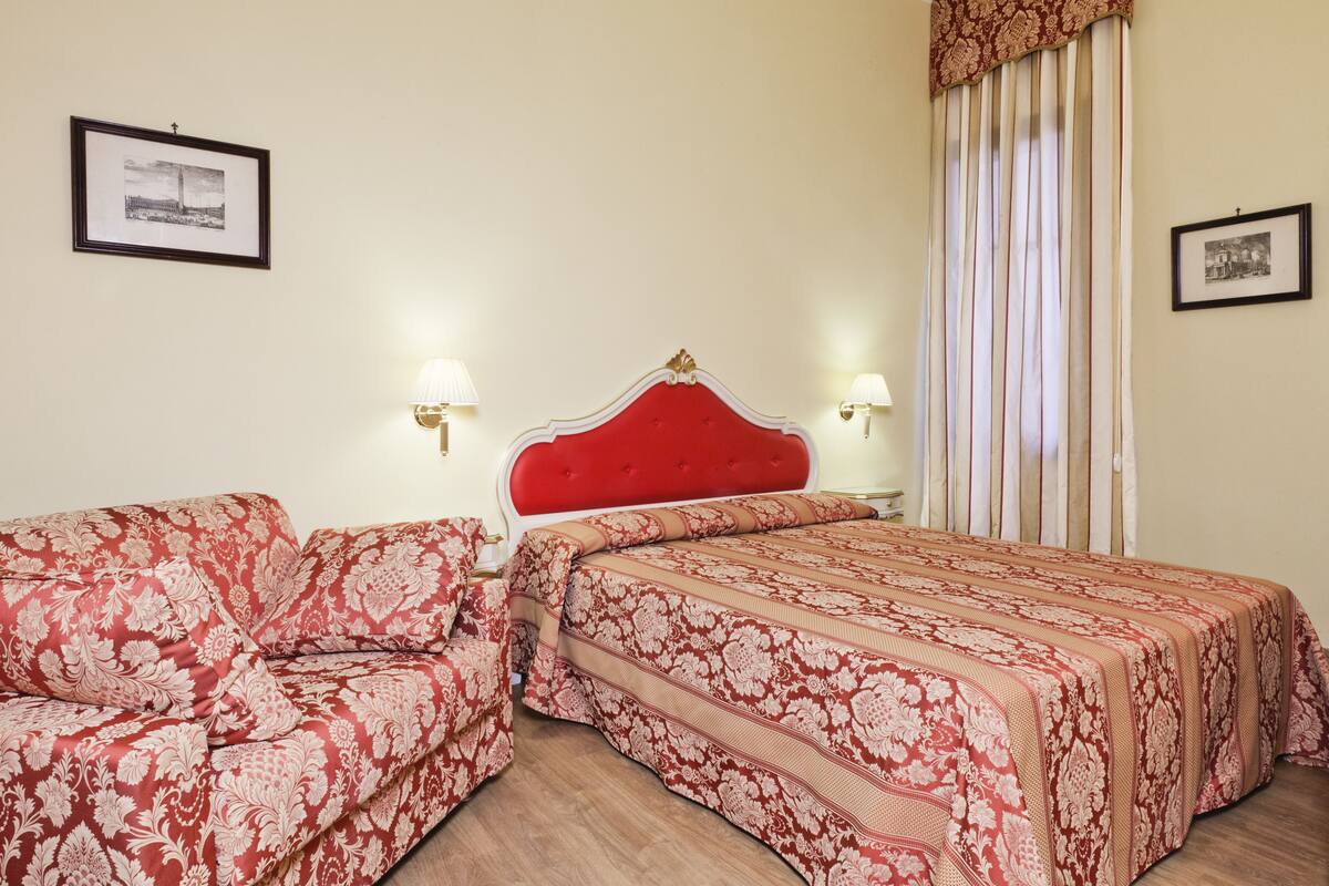 A hotel room with a red upholstered bed, a matching sofa, and two wall-mounted lamps.
