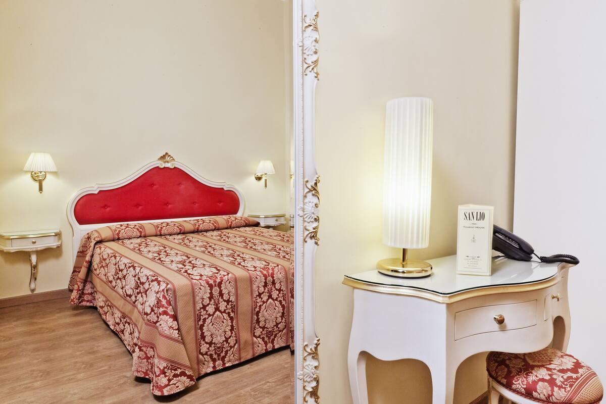 A hotel room with a red upholstered headboard, a bed with a floral-patterned bedspread, a bedside table with a lamp, and a desk with a telephone and a sign that reads "SAN LIO TOURIST HOUSE".