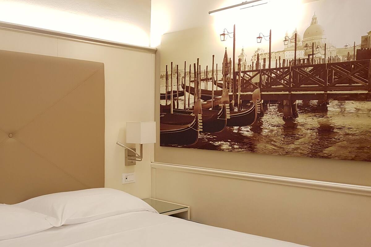 A hotel room with a bed, a wall-mounted lamp, and a large painting of a Venetian canal scene.