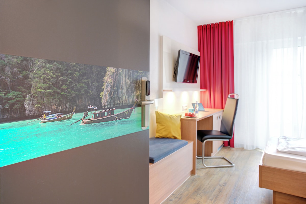 Habitación de hotel con cama, escritorio, silla, televisor y un cuadro de barcos en el agua.