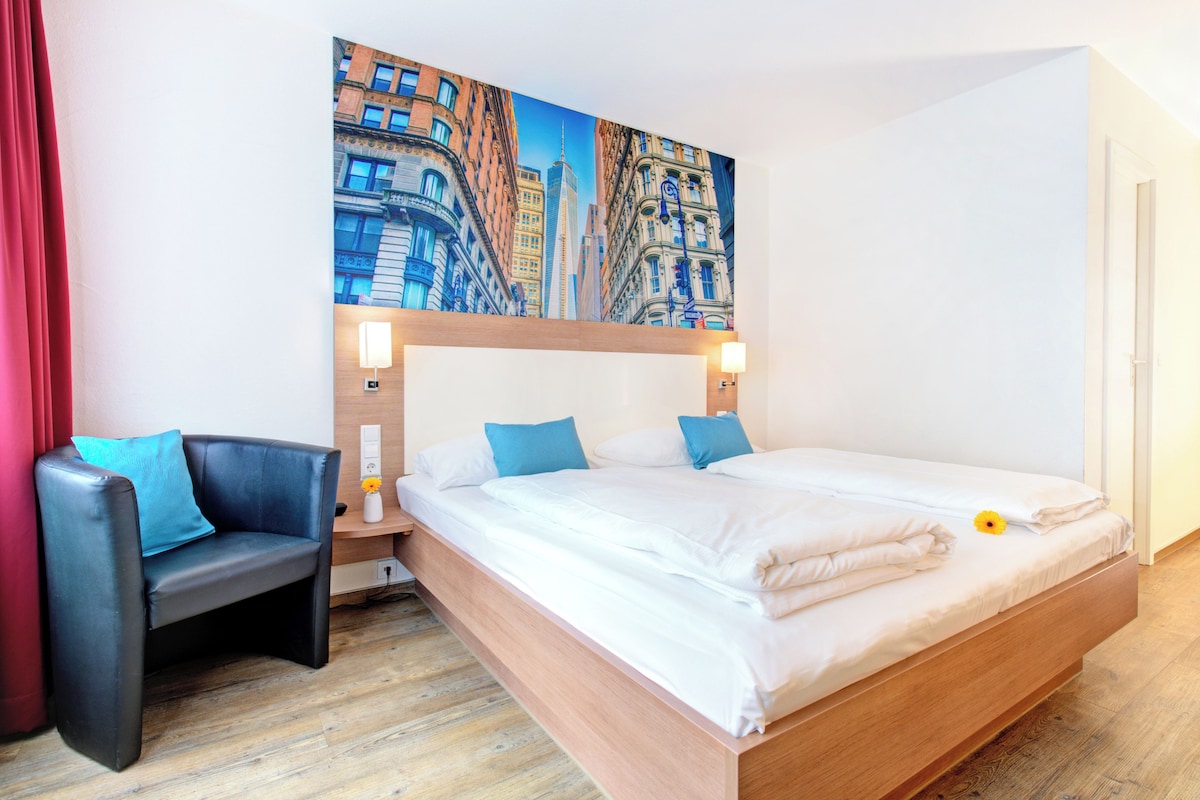 Habitación de hotel moderna con una cama grande, una mesita de noche, una silla y un cuadro con una vista urbana.