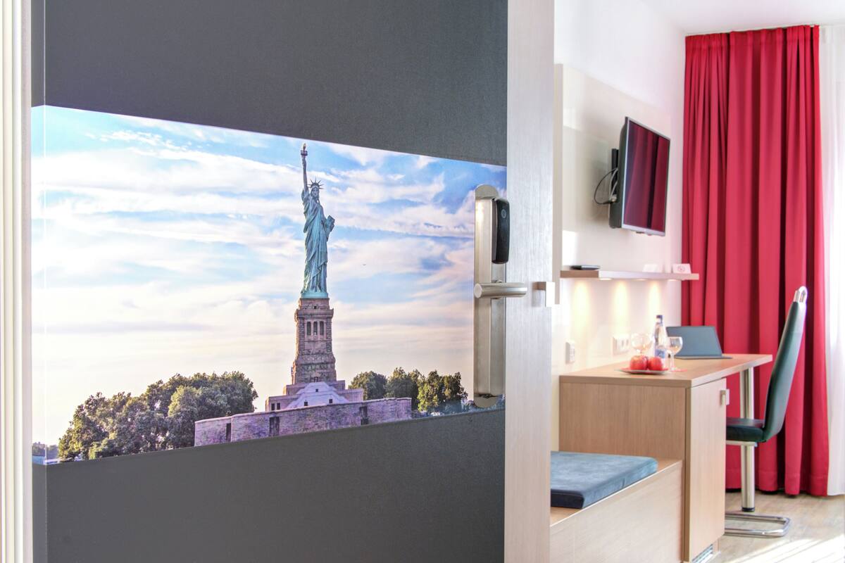 Ein Hotelzimmer mit großem Fenster, das die Freiheitsstatue zeigt, einem Schreibtisch mit einem Laptop, einem Sessel und einem an der Wand montierten Fernseher.