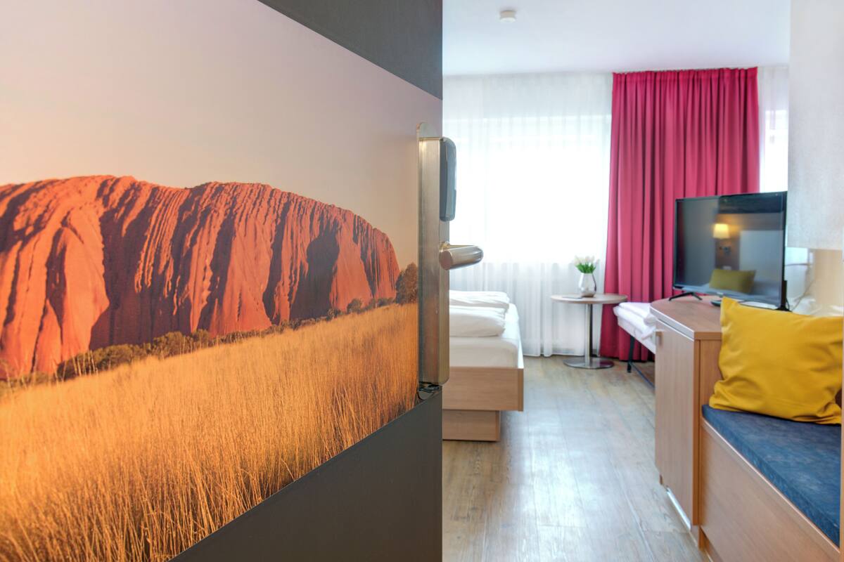 Ein Hotelzimmer mit einer großen Wandmalerei einer Wüstenlandschaft, einem Bett mit einem gelben Kissen, einem Fernseher und einem kleinen Tisch mit einer Pflanze.