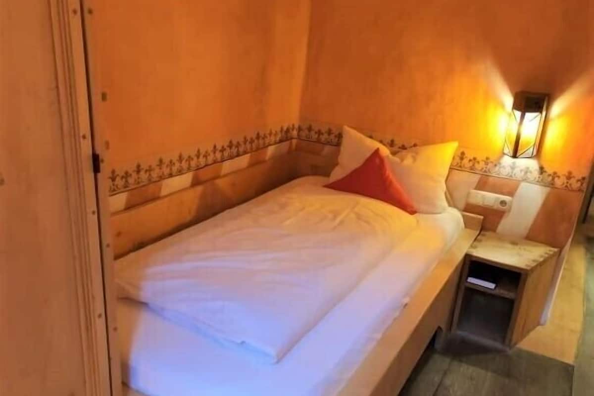A wooden bed with white bedding and a red pillow in a room with wooden walls and a nightstand.