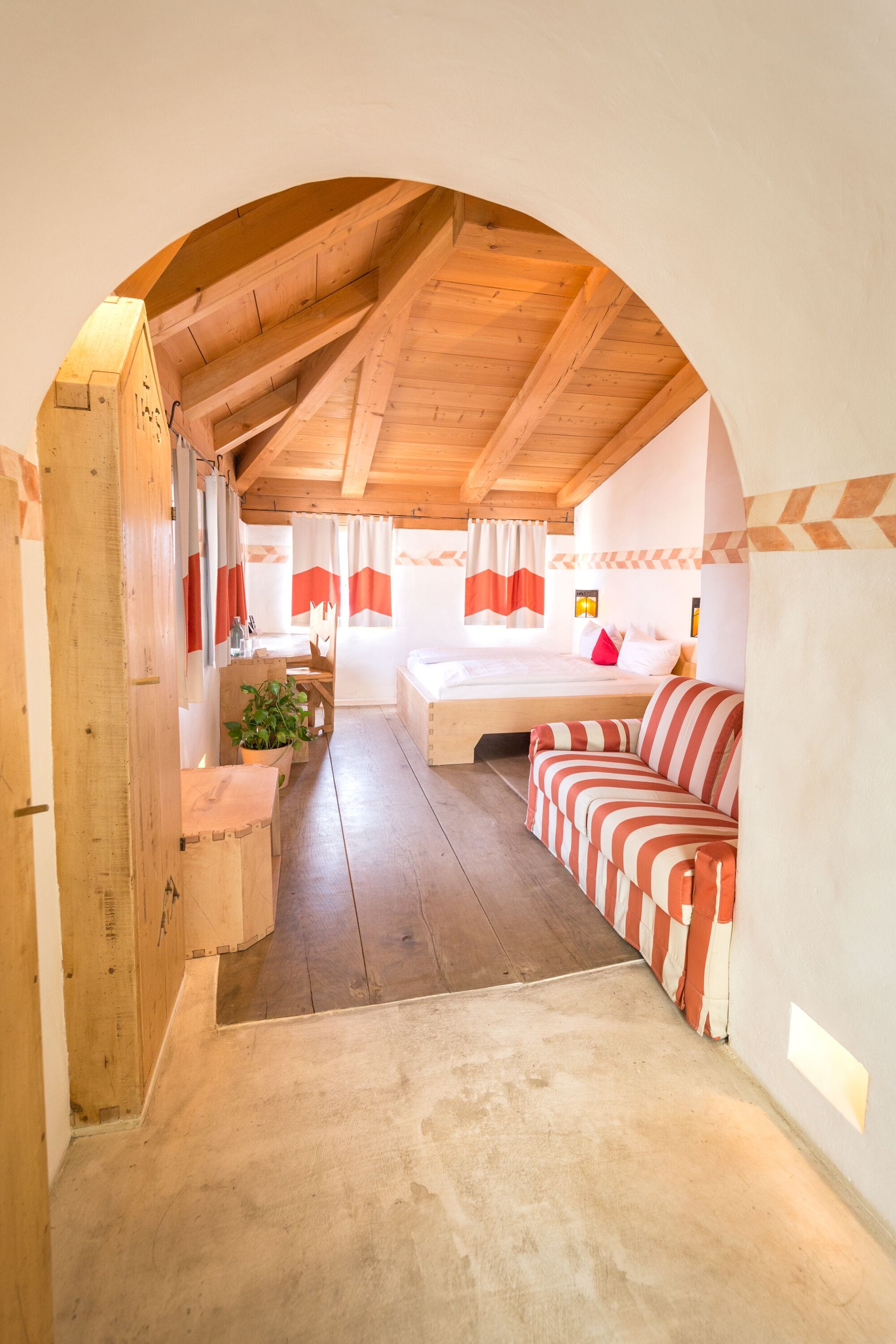 Ein gemütliches Zimmer mit Holzbalkendecke, einem Bett mit rot-weißer Bettwäsche, einer gestreiften Couch und einem kleinen Tisch mit einer Pflanze.