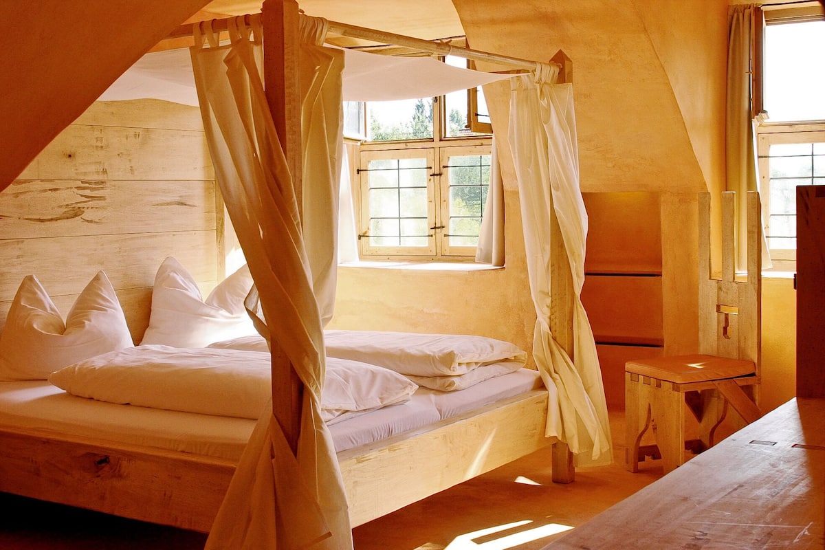 A wooden room with a bed, a small table, and a window with curtains.