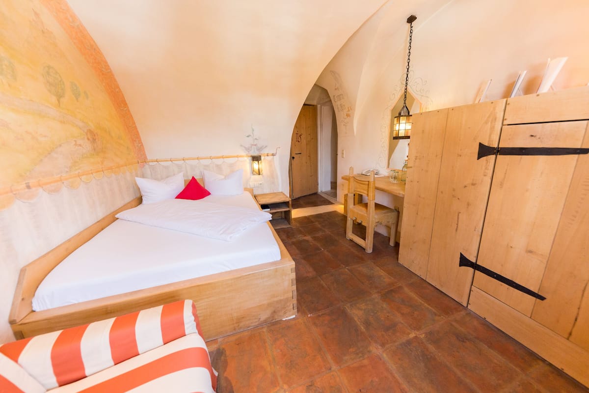 A cozy bedroom with a wooden bed, a desk, and a chair, featuring a rustic ceiling and wooden walls.