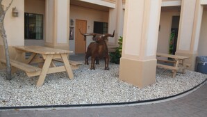 Un patio con dos mesas de picnic de madera, una estatua de toro y un edificio de paredes color beige y ventanas.