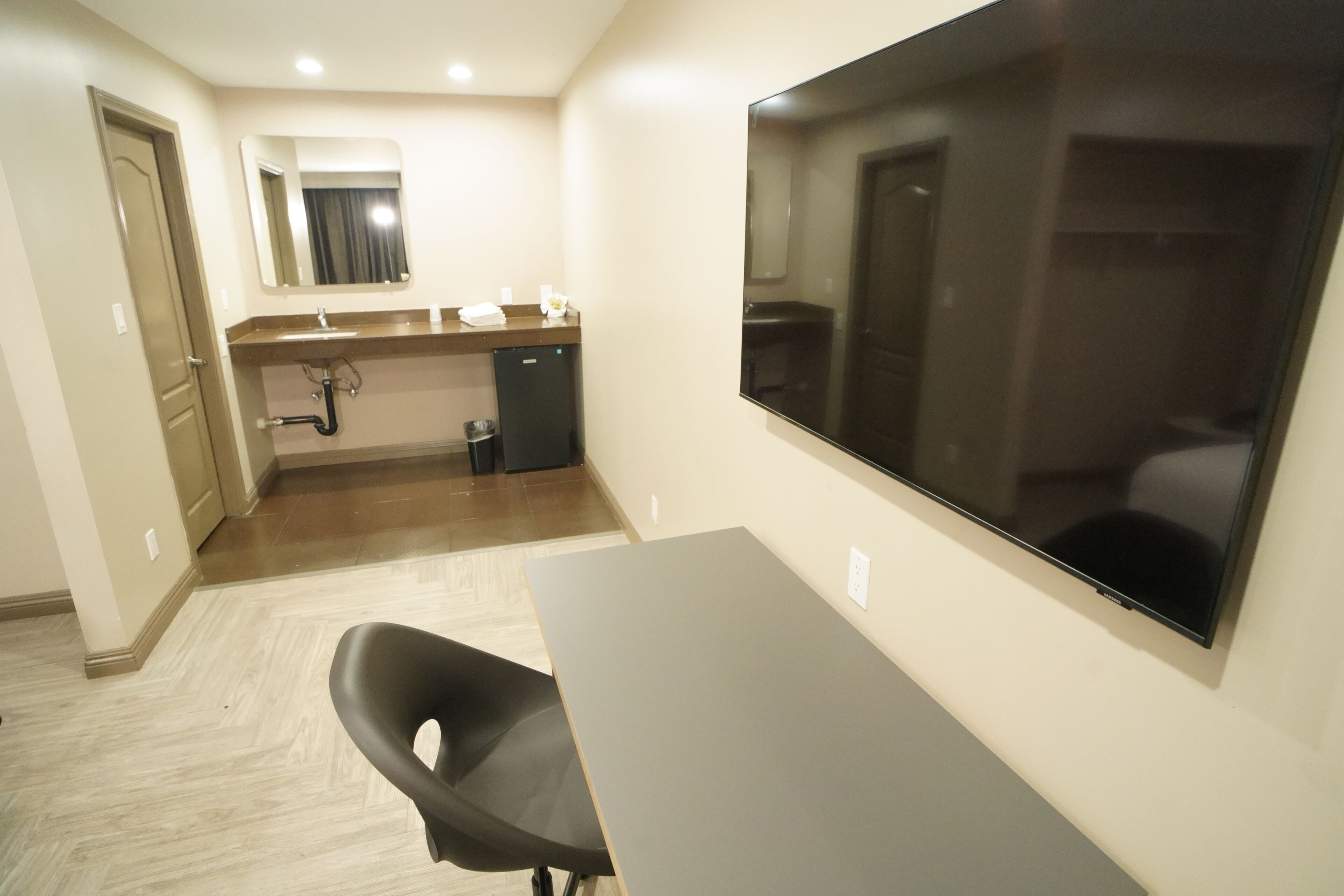A hotel room with a large flat-screen TV, a desk, a chair, a small kitchenette, and a mirror.