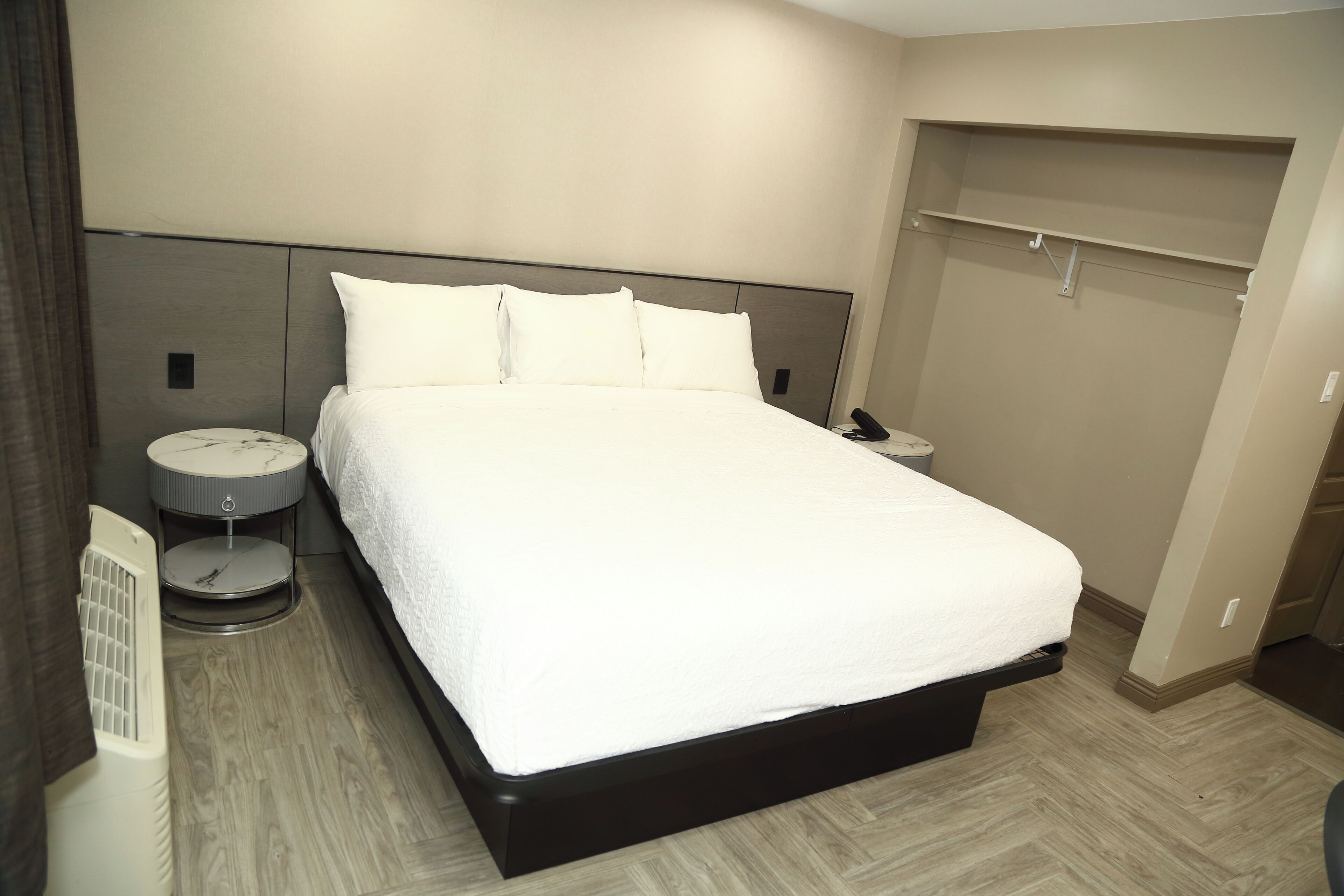 A modern hotel room with a large bed, bedside tables, a round nightstand, a built-in closet, and a wall-mounted heater.