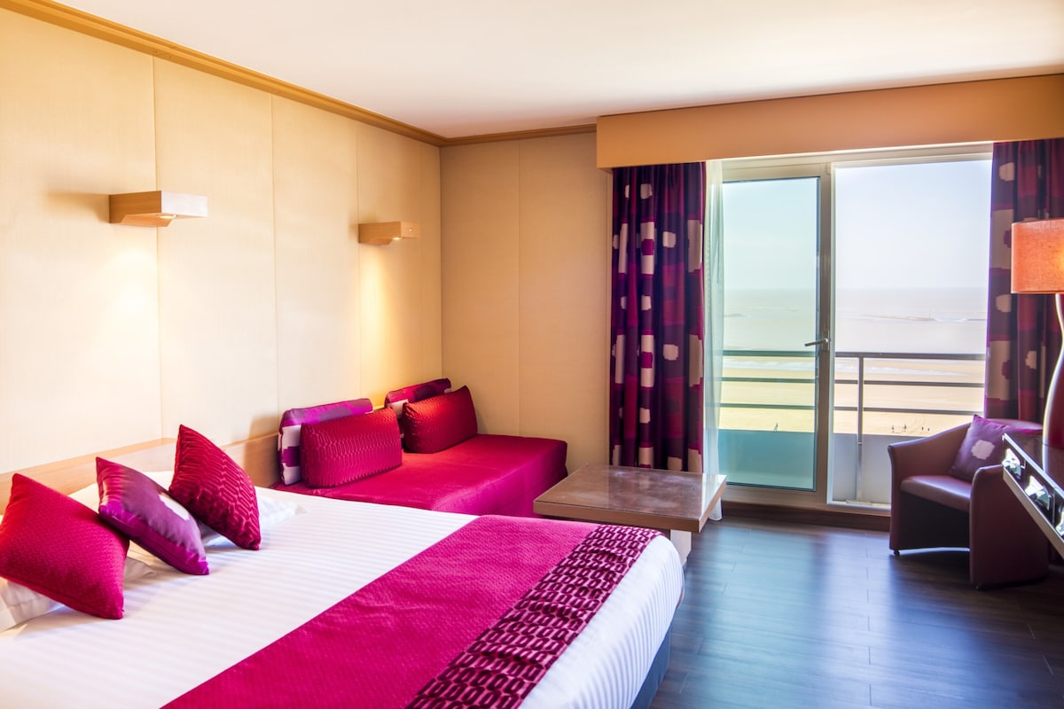 Habitación de hotel con dos camas, un sofá, una mesa y un balcón con cortinas.