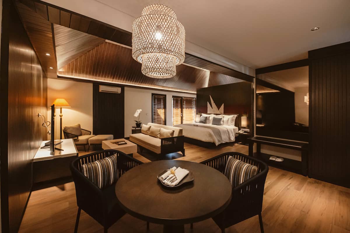Una habitación de hotel moderna con techo de madera, una cama grande, una zona de estar con sofá y sillones, una mesa de comedor y una lámpara de araña grande.