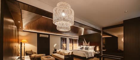 A modern hotel room with a wooden ceiling, a large bed, a sitting area with a sofa and armchairs, a dining table, and a large chandelier.