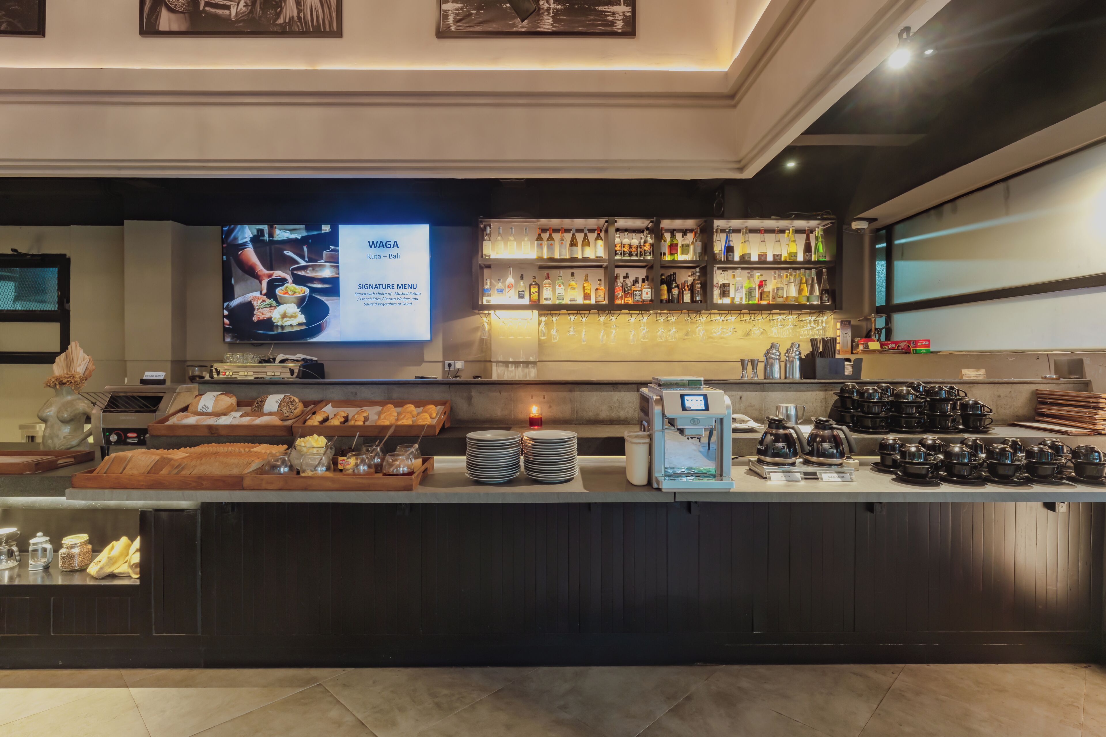 A hotel bar area with a digital menu screen, a variety of beverages, and a coffee machine.