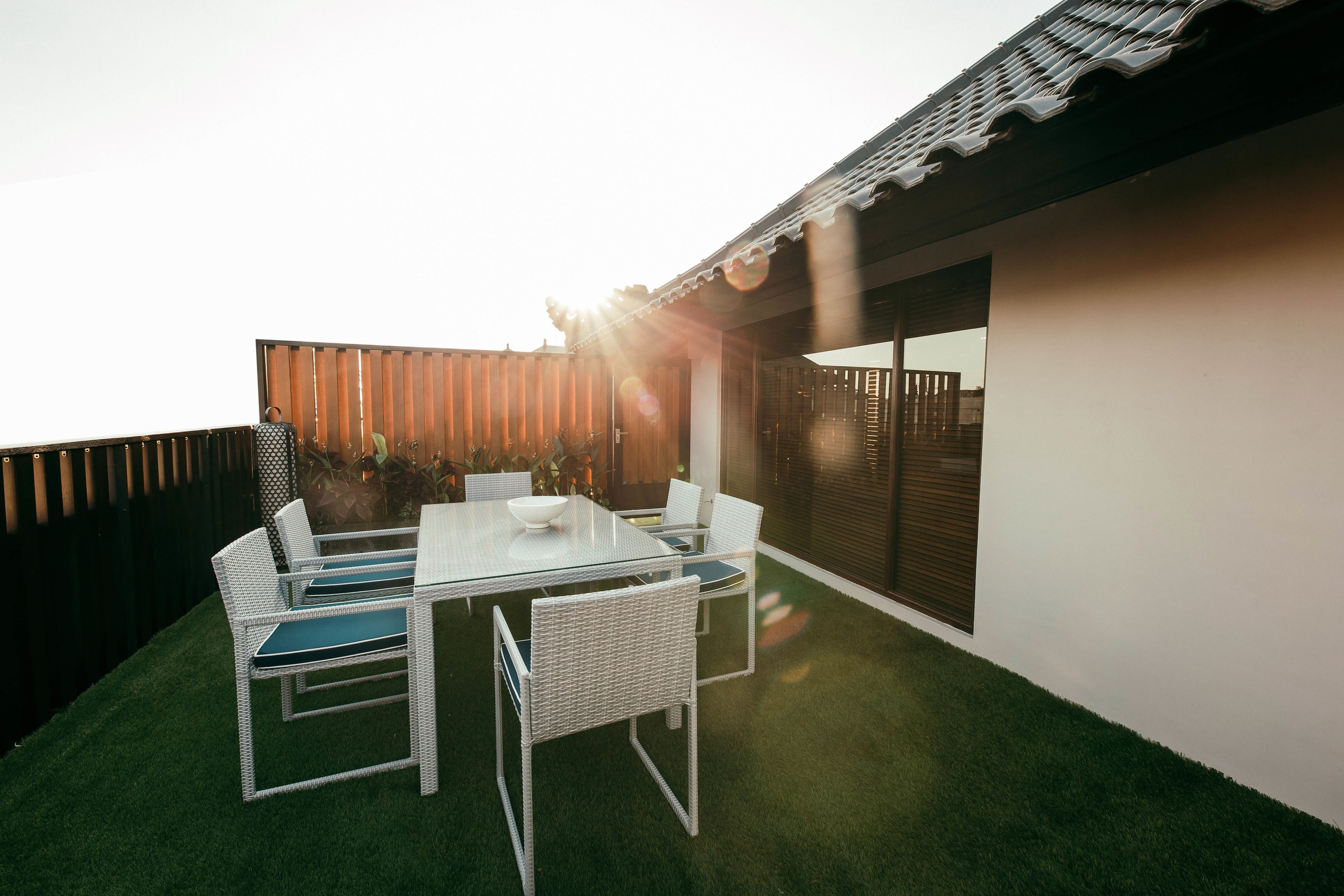 An outdoor patio with a glass table and white wicker chairs, surrounded by artificial grass.