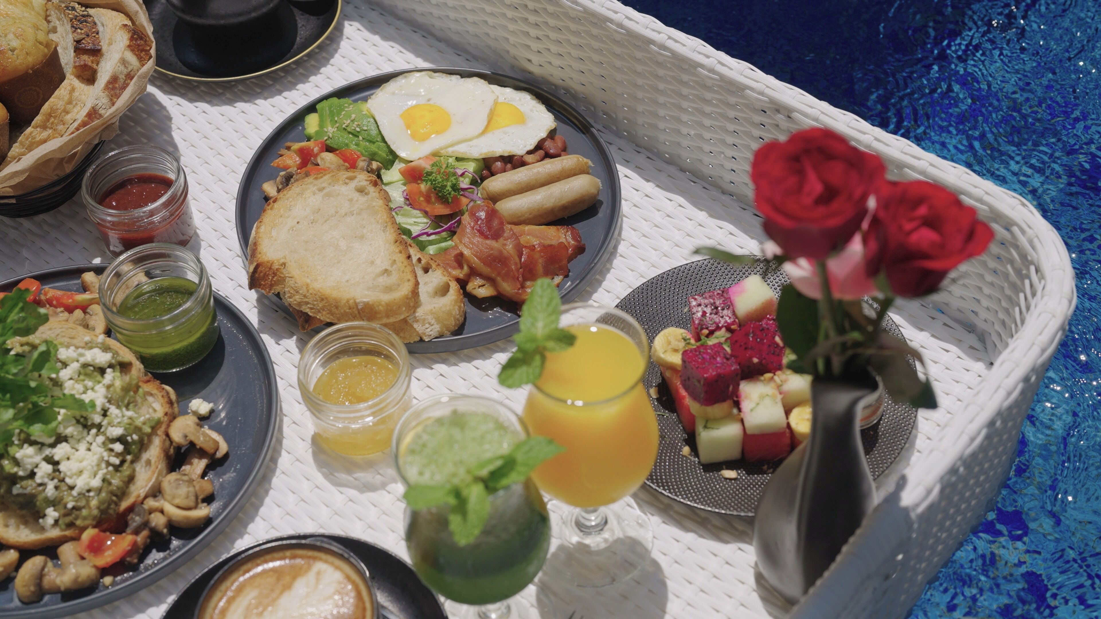 Bandeja de desayuno con tostadas, huevos, tocino y diferentes acompañamientos, incluyendo fruta y una bebida, sobre una mesa junto a una piscina.