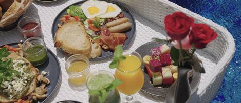 Bandeja de café da manhã com torradas, ovos, bacon e acompanhamentos diversos, incluindo frutas e uma bebida, sobre uma mesa perto de uma piscina.