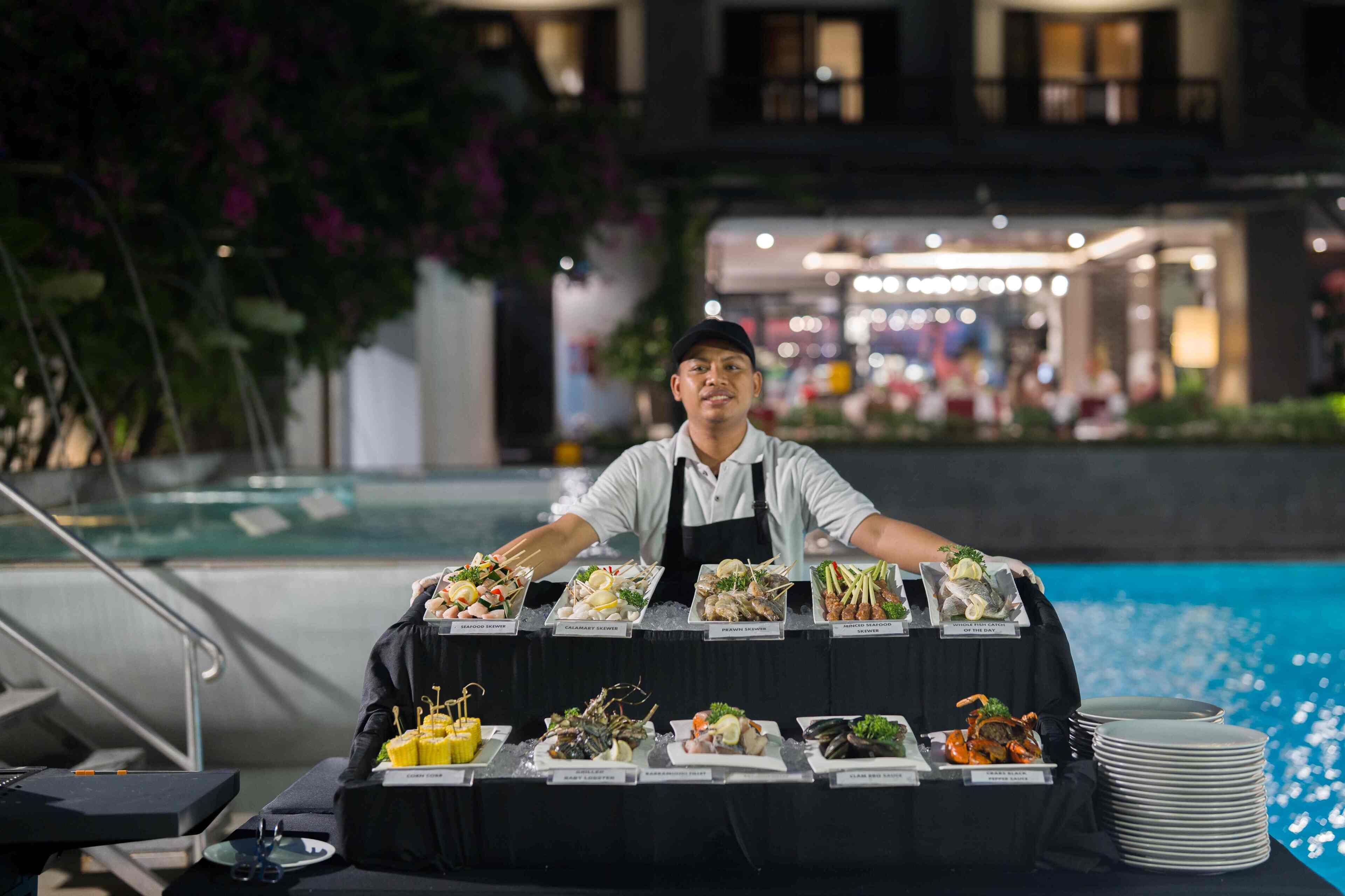 A buffet table with various seafood dishes, a person serving, and a pool in the background.