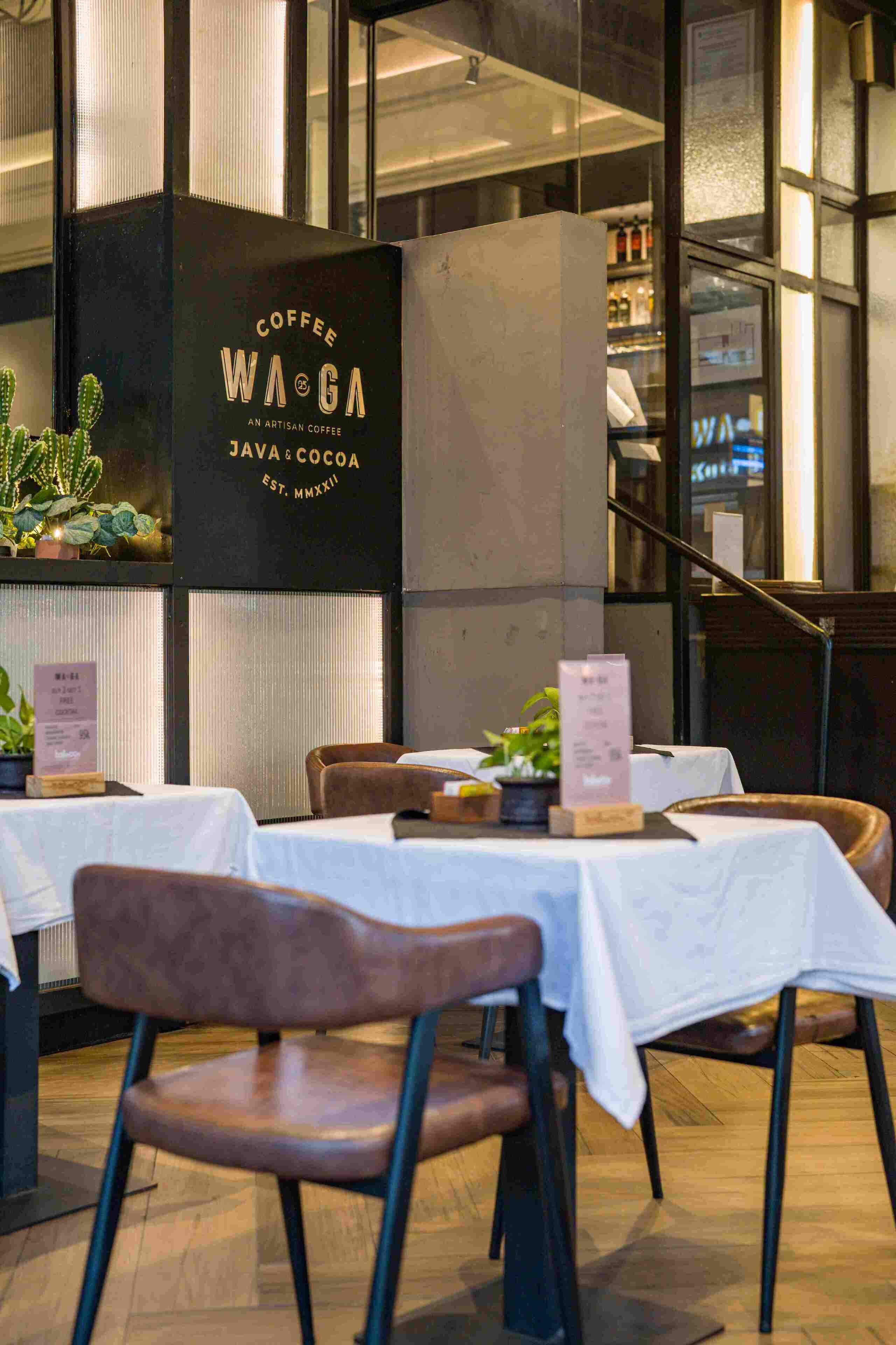 Interior de una cafetería con mesas y sillas, un letrero con el nombre del establecimiento, y una vista del exterior a través de amplios ventanales.