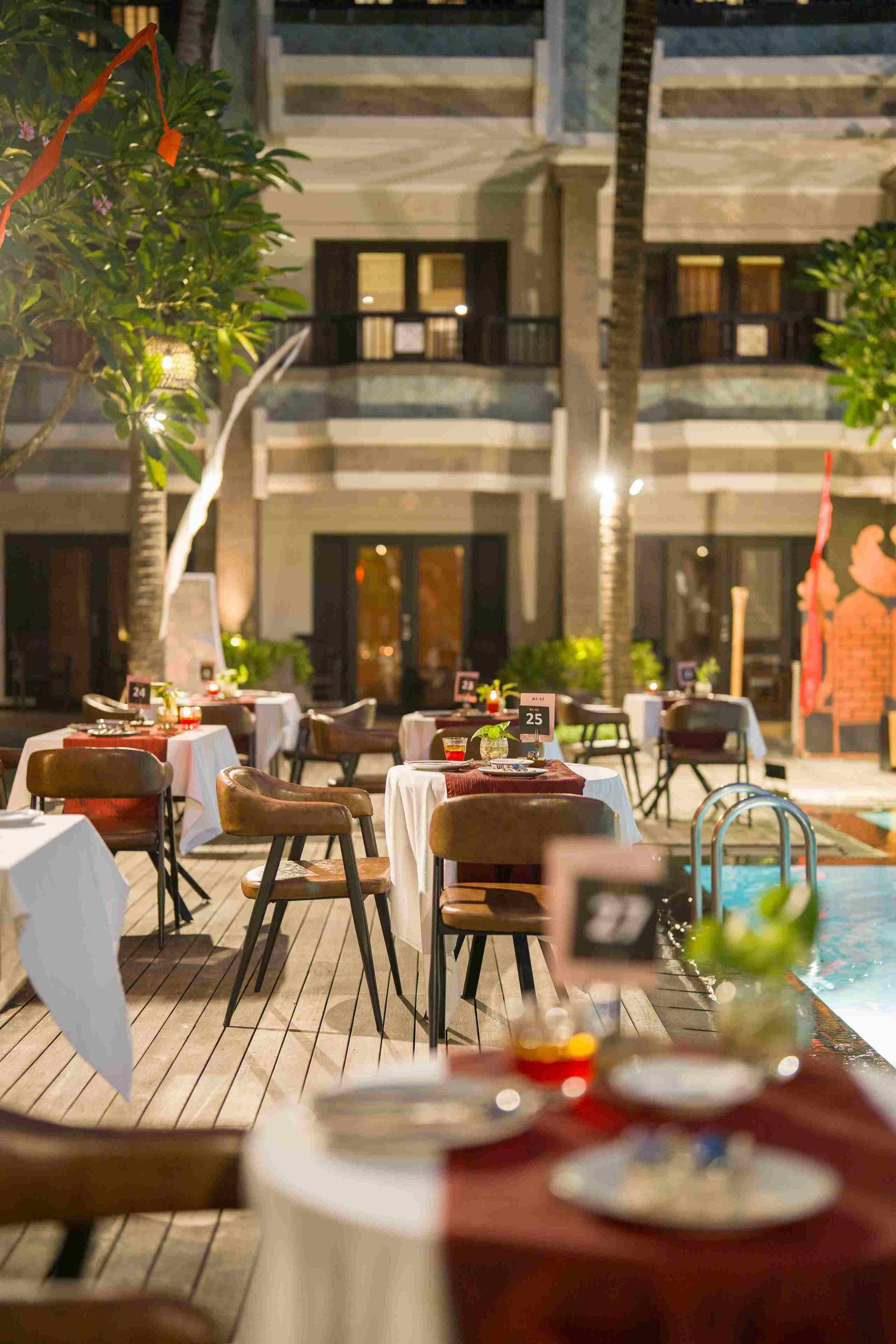 Un área de comedor al aire libre con mesas y sillas, una piscina y un edificio al fondo.