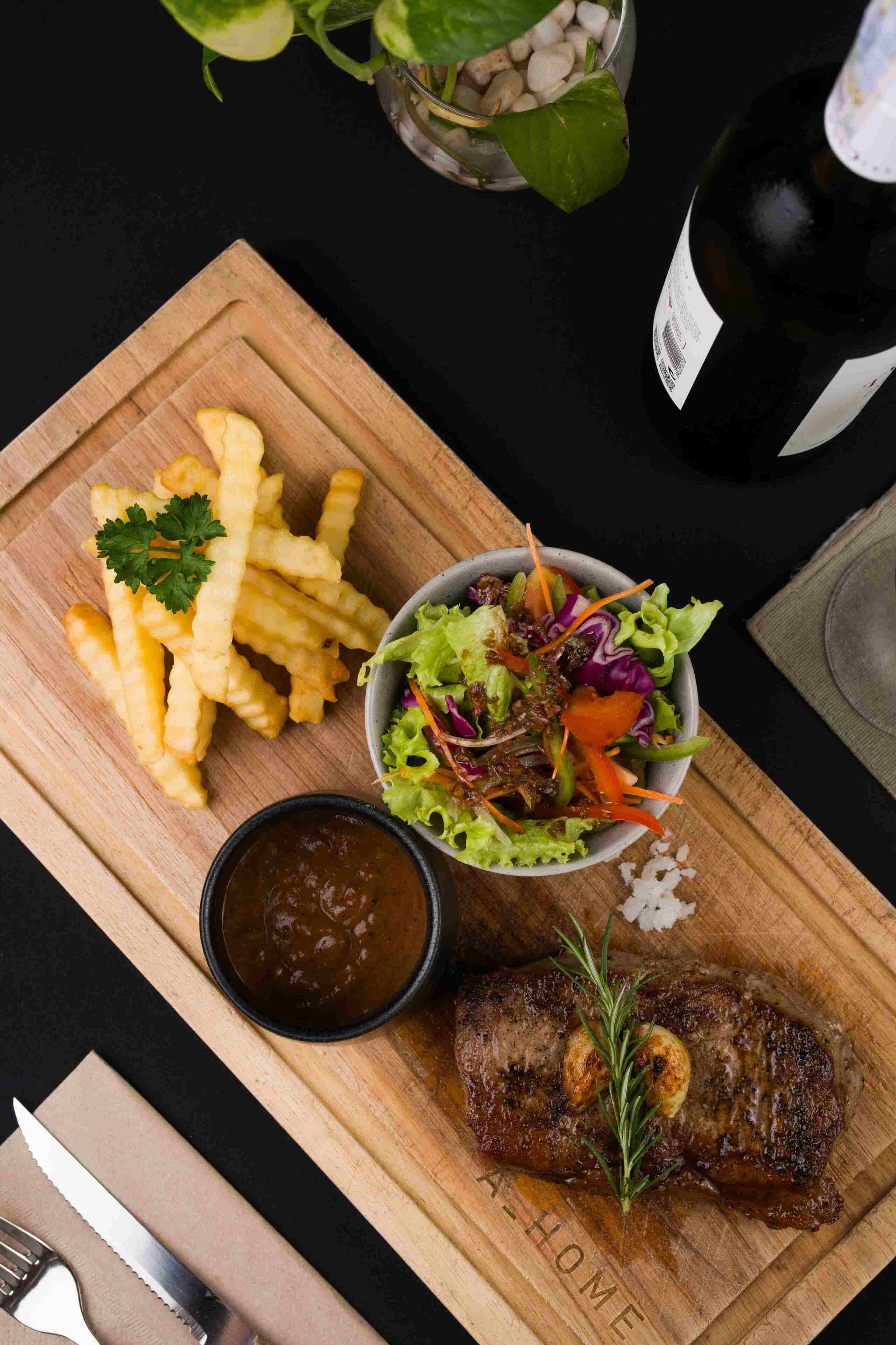 A wooden cutting board with a meal consisting of fries, a salad, a steak, and a sauce.