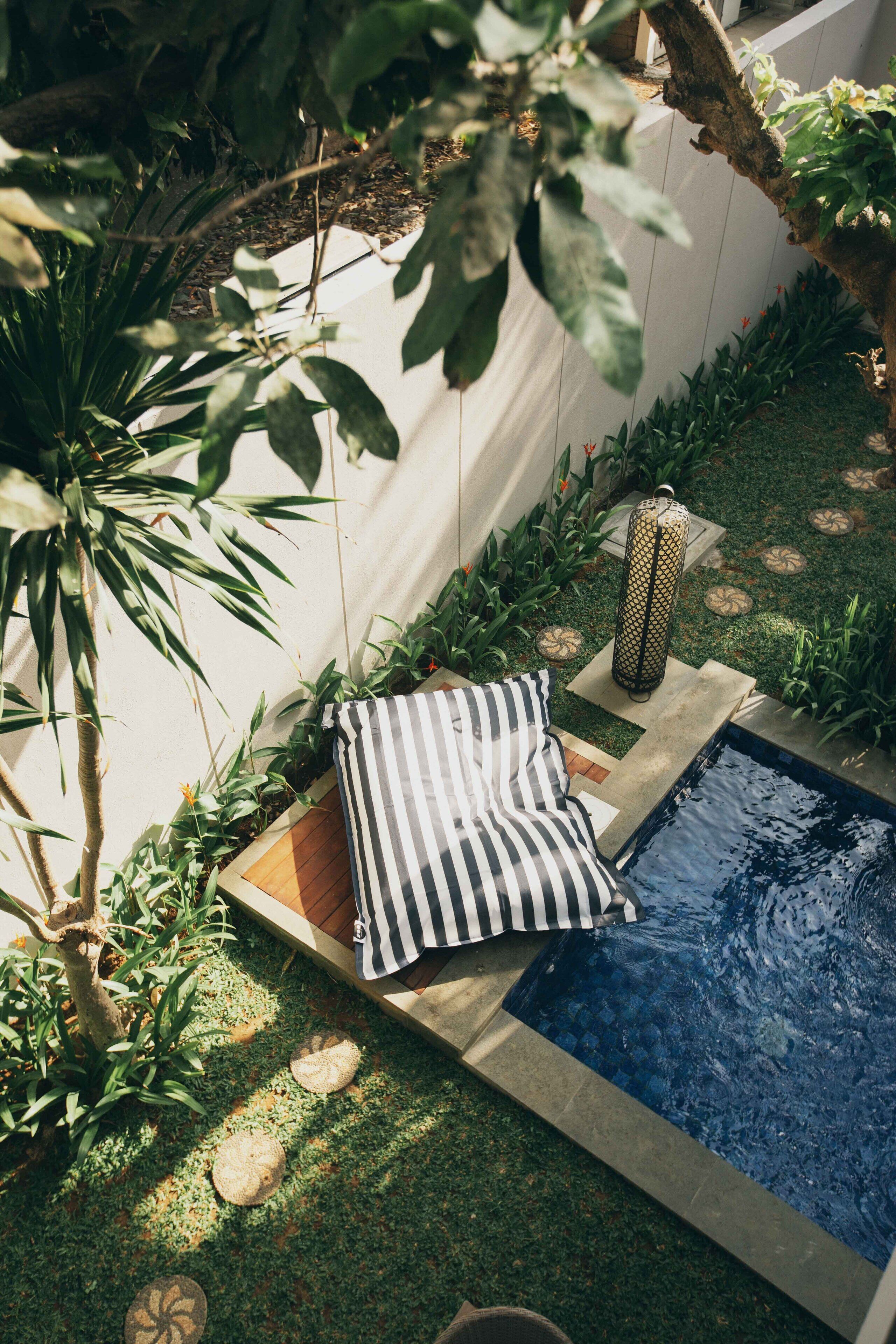 A small pool area with a wooden deck, a striped cushion, and surrounding greenery.