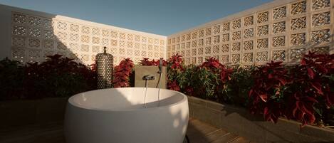 Una bañera de hidromasaje circular en una terraza de madera, con una cerca decorativa y plantas al fondo.