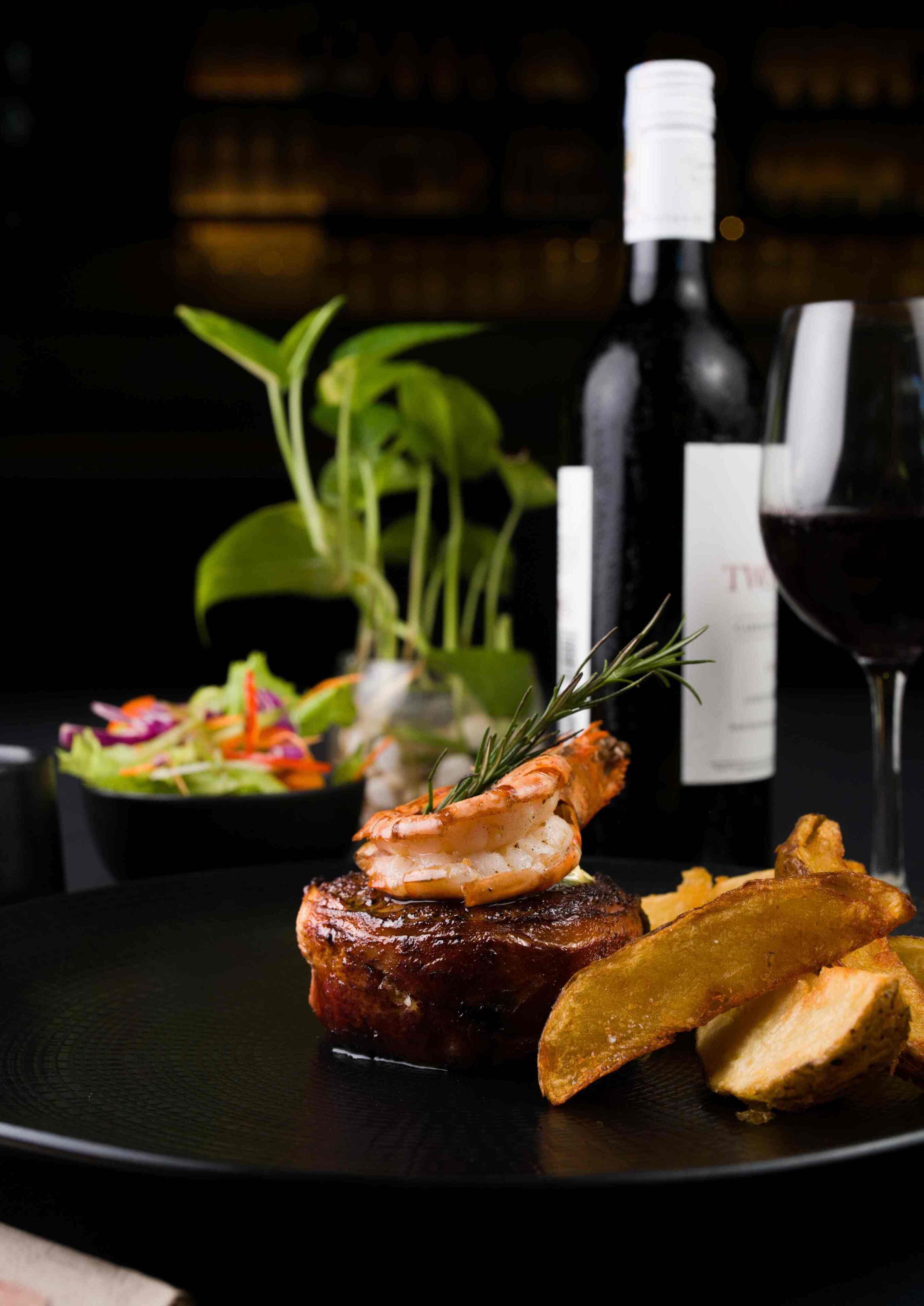 Une assiette contenant un steak, des crevettes et des quartiers de pommes de terre, un verre de vin rouge et une bouteille de vin sur une table sombre.