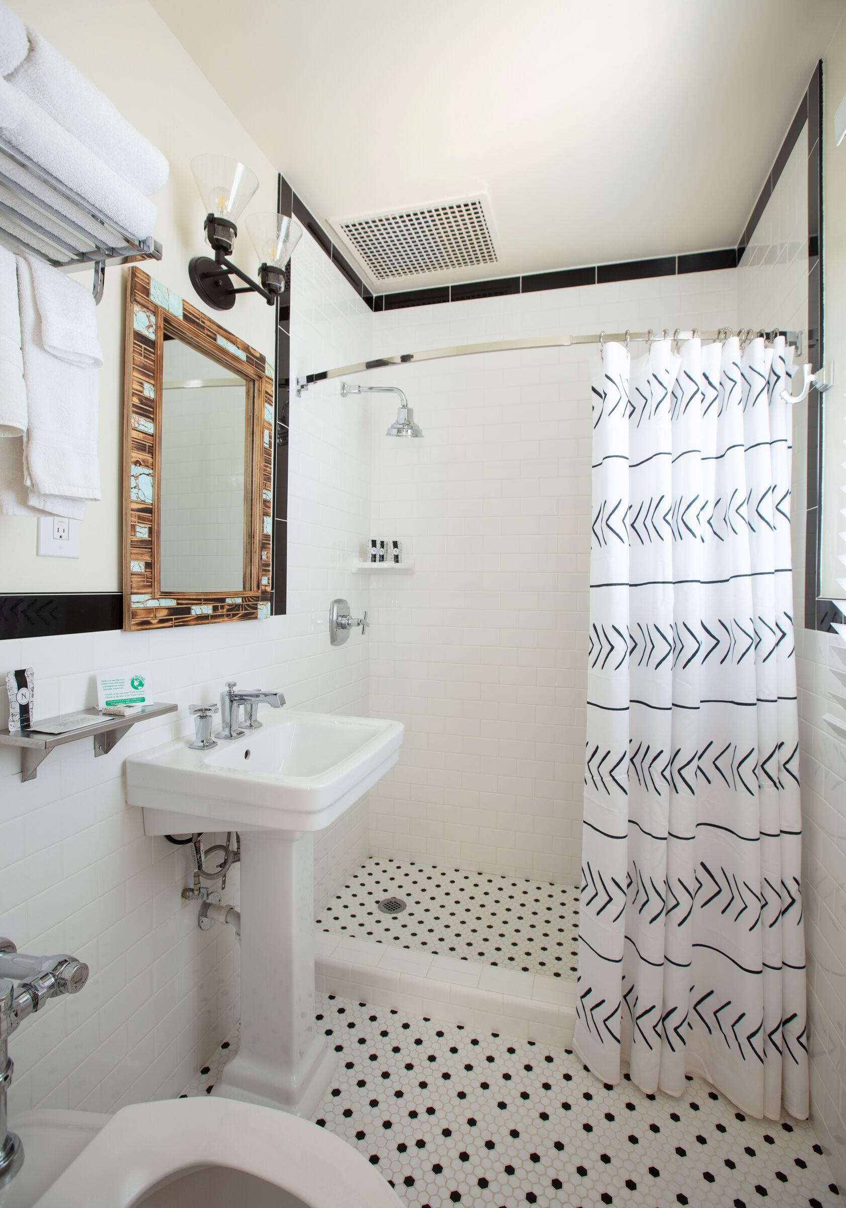 Un baño con un lavamanos blanco, un espejo, una cortina de ducha con diseño geométrico y un suelo de baldosas con diseño blanco y negro.
