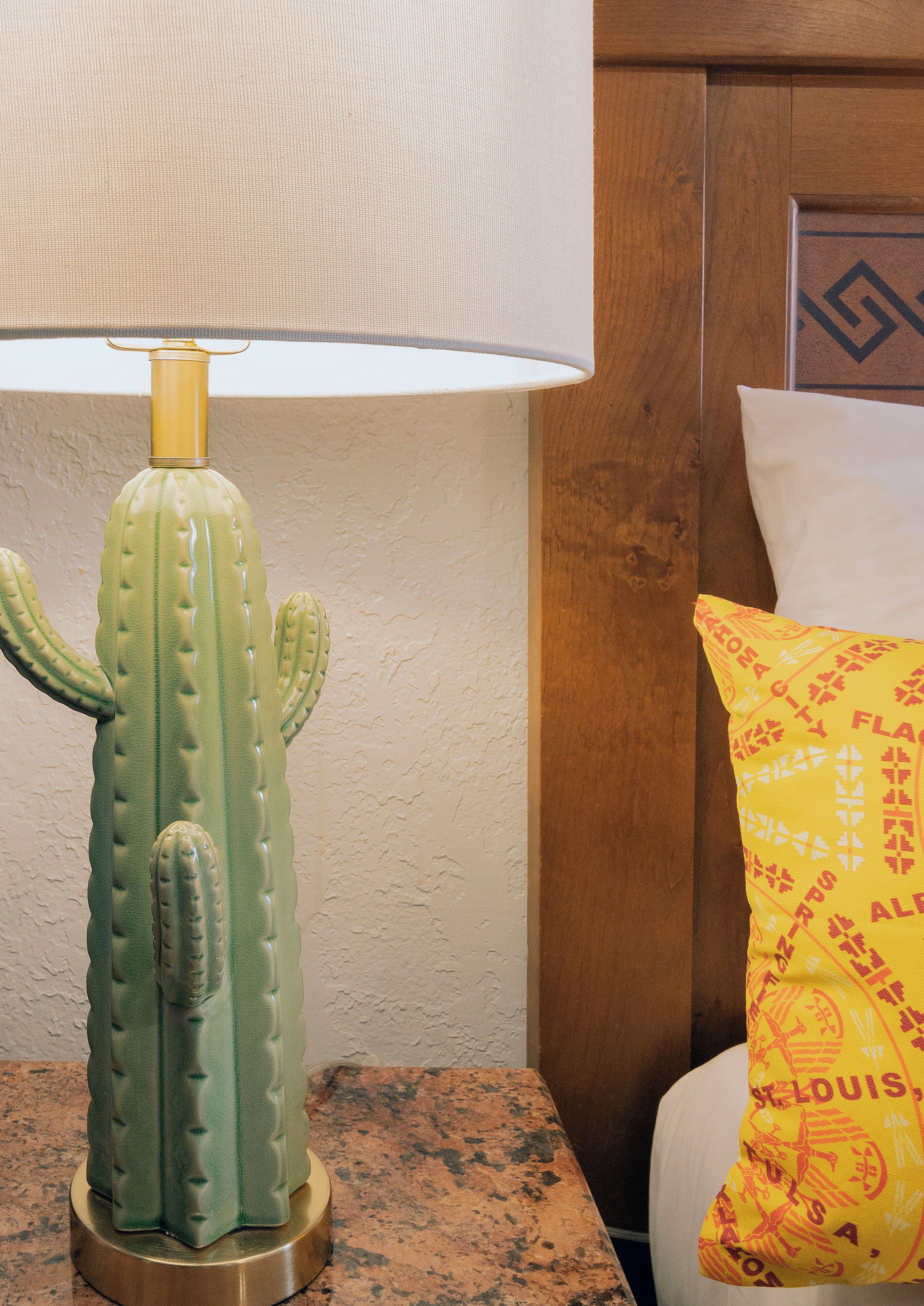 A cactus-shaped lamp on a table next to a bed with a patterned pillow.