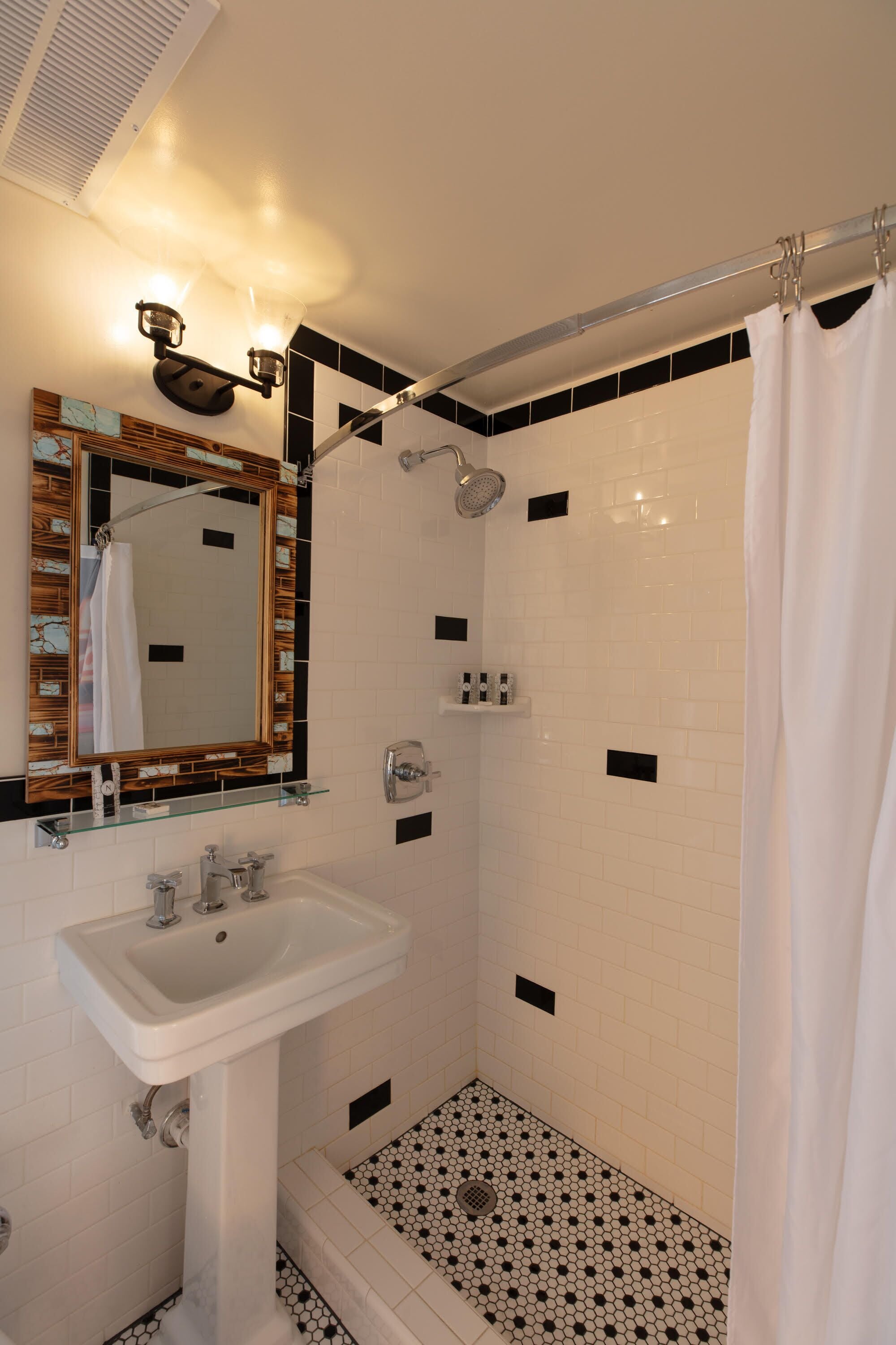 A bathroom with a white sink, a mirror with a decorative frame, a shower with a curtain, and a tiled floor with a black and white pattern.
