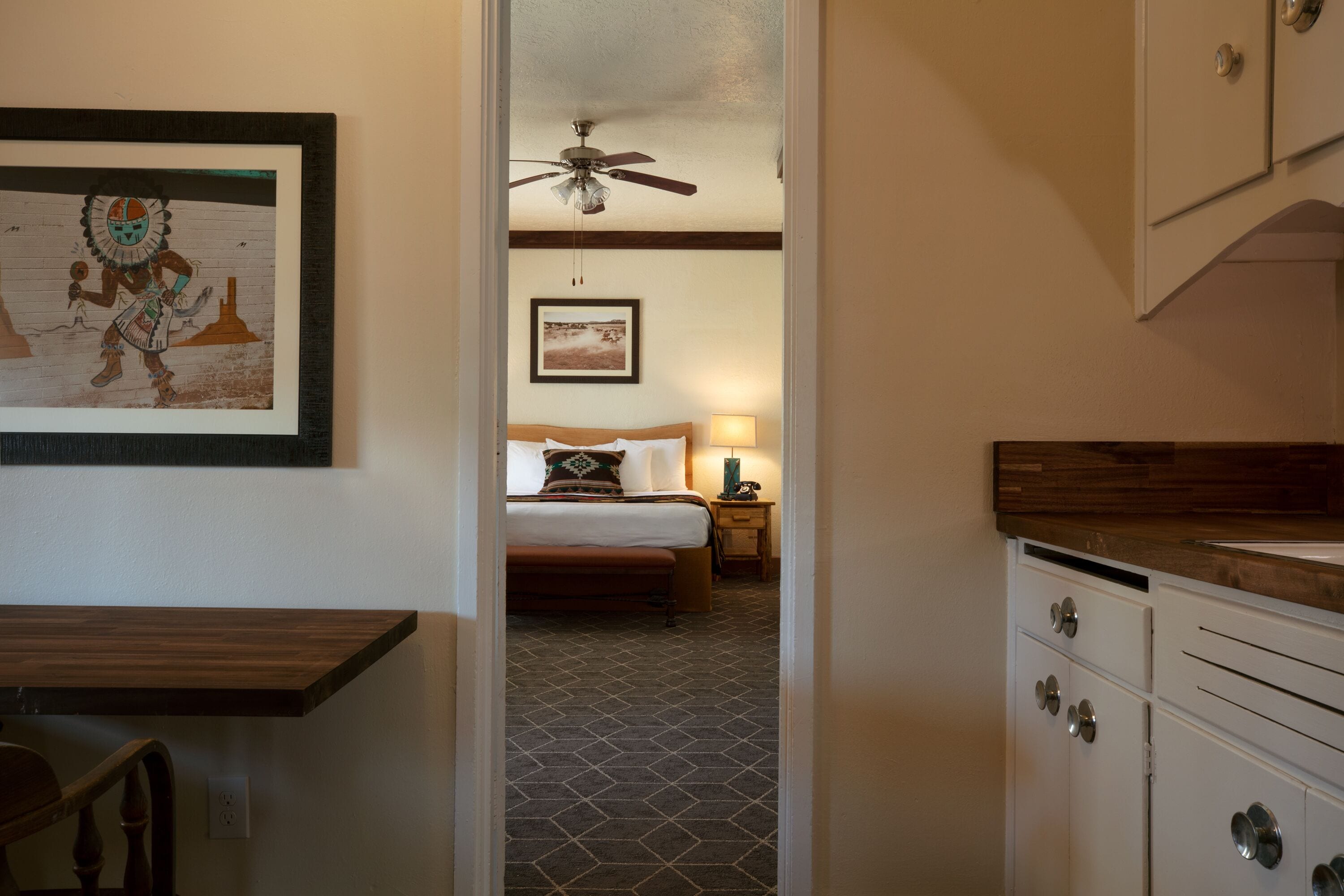 A hotel room with a bed, a nightstand, a ceiling fan, and a kitchen area with cabinets.
