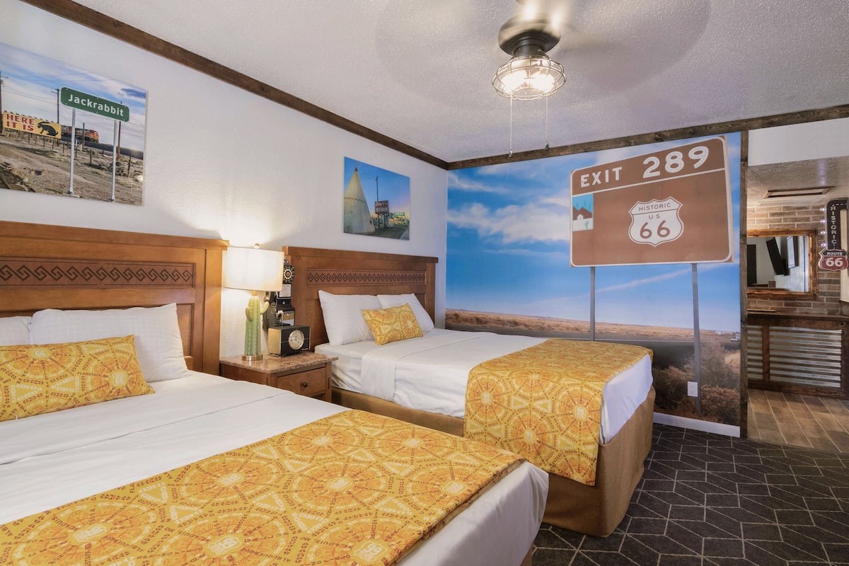 A hotel room with two beds, a nightstand, a lamp, a cactus, and a mural of a desert landscape with a sign for "Exit 289 Historic US 66".