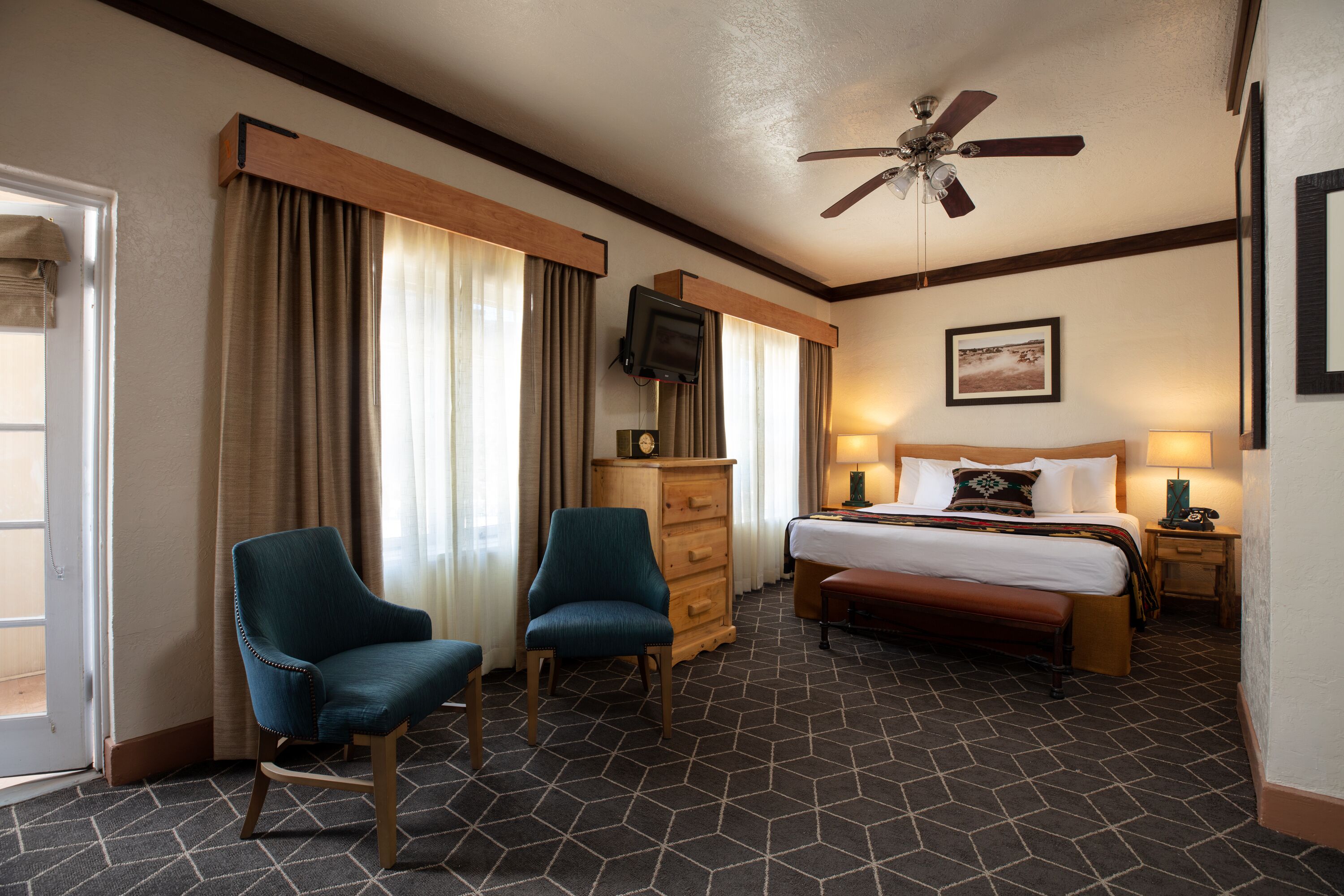 A hotel room with a bed, two chairs, a dresser, a television, and a ceiling fan.