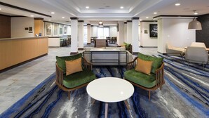 A hotel lobby with a reception desk, a seating area with two green upholstered chairs and a white table, and a dining area in the background. - Fairfield Inn & Suites by Marriott Elizabethtown (Elizabethtown)