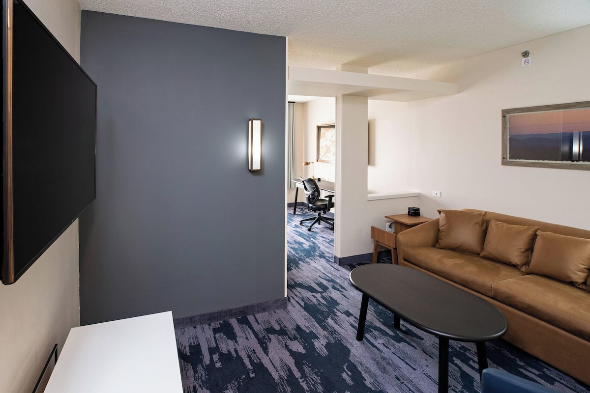 A hotel room with a dark grey wall, a brown leather sofa, a small table, and a TV mounted on the wall.
