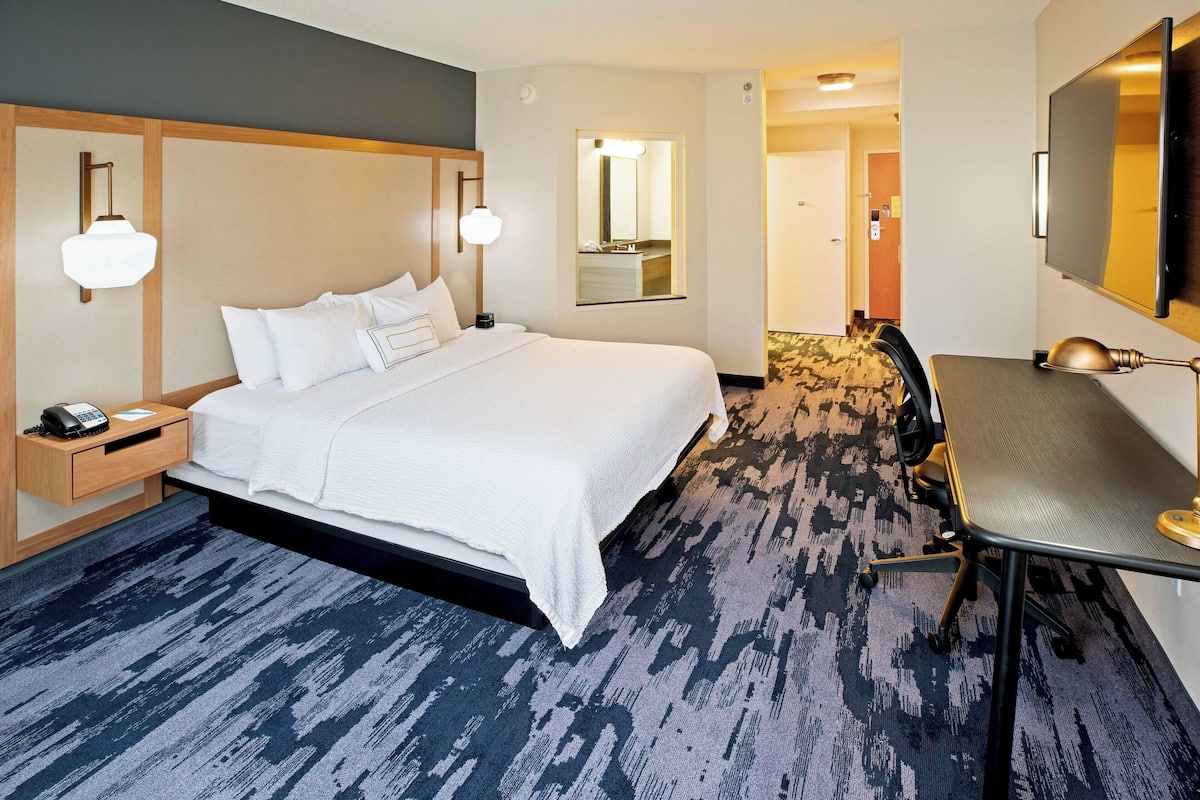 A hotel room with a large bed, bedside table with a lamp, a desk with a chair, and a large mirror on the wall.