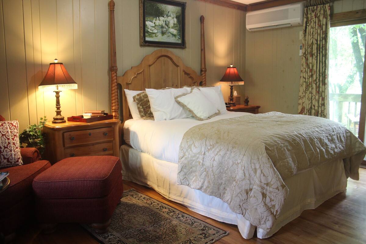 A bedroom with a large bed, two bedside tables with lamps, a wooden headboard, a red armchair, and a patterned rug.
