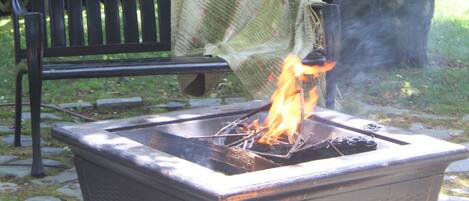 A fire pit with flames, a bench, and a wooden deck in a garden setting.