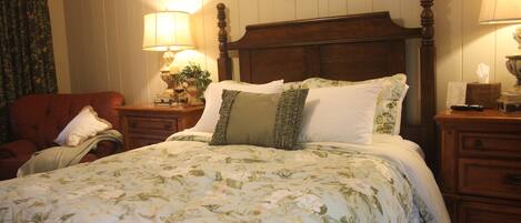 A neatly made bed with floral bedding, a wooden headboard, and a bedside table with a lamp and a framed picture on the wall.