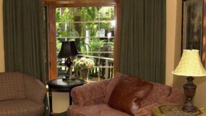 A living room with a brown leather sofa, a matching ottoman, a small round table with a lamp, and a view of greenery through a window with curtains. - The Village Inn (Blowing Rock)
