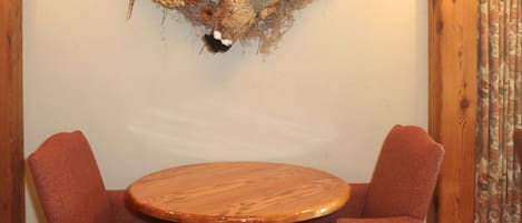 A wooden dining table with two red chairs, a wooden wall with a decorative wreath, and a wooden-framed window with a lamp.