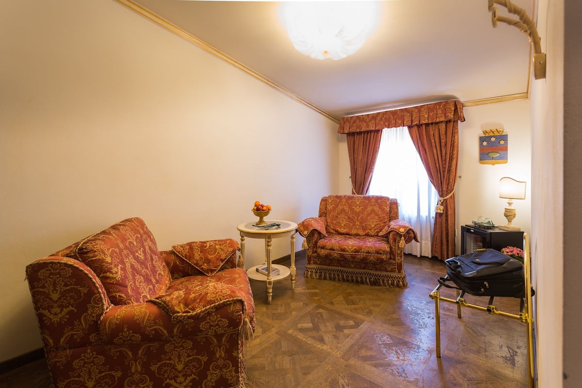 A room with a patterned sofa, a matching armchair, a small round table with a fruit bowl, and a suitcase on a stand.