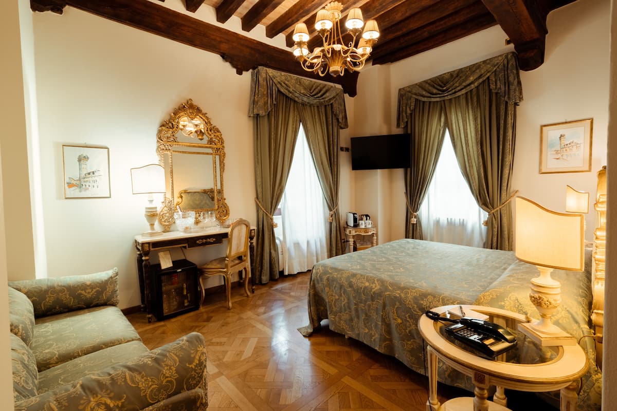 A hotel room with a large bed, a desk with a chair, a television, and a chandelier.