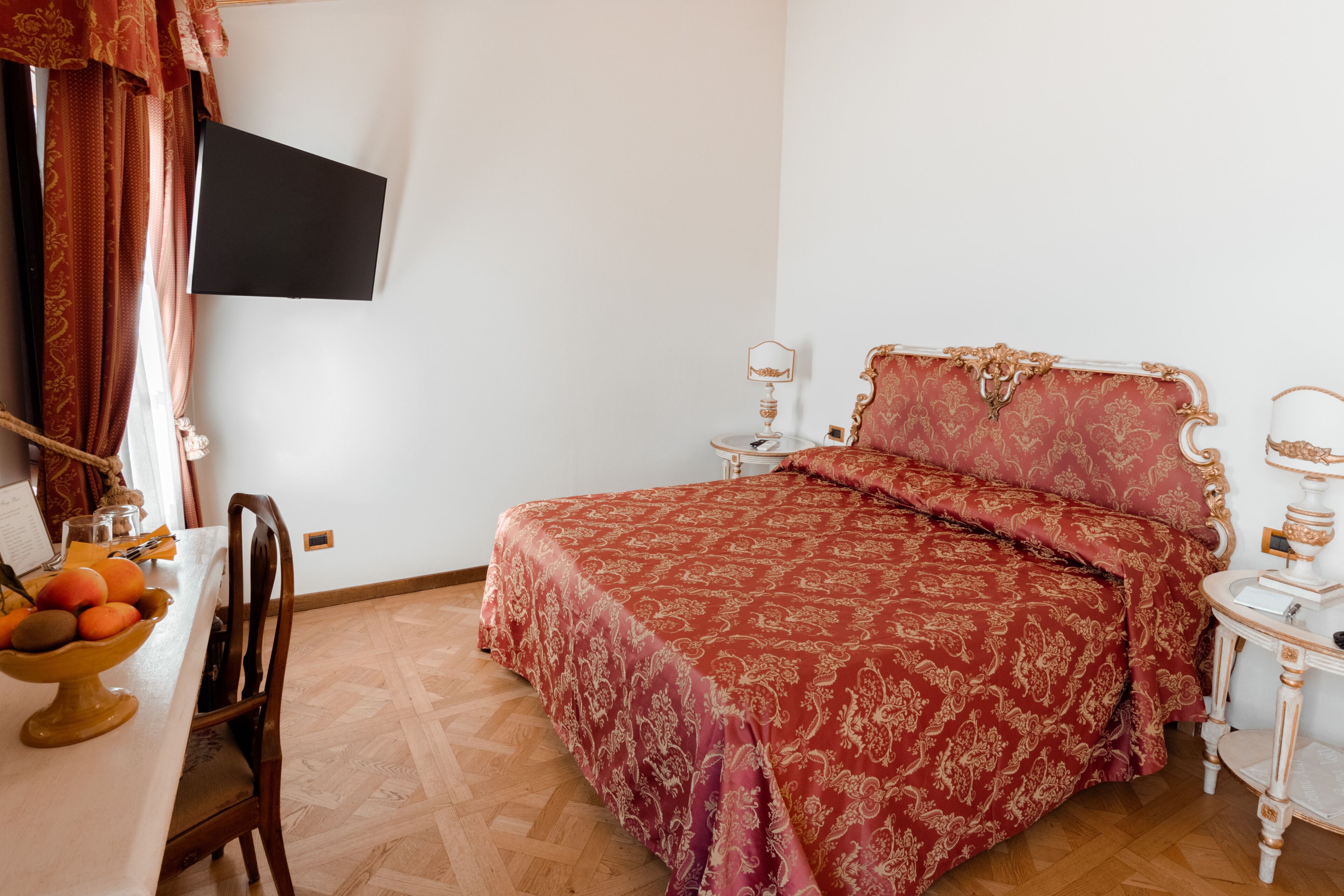 Une chambre avec un grand lit, un bureau sur lequel se trouve un panier de fruits, une chaise, un téléviseur fixé au mur et une table de chevet avec une lampe.