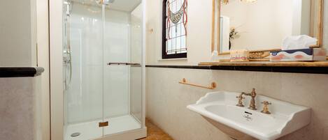Un baño con ducha envuelta en vidrio, un lavamanos blanco con un espejo decorativo y una ventana con vitrales.