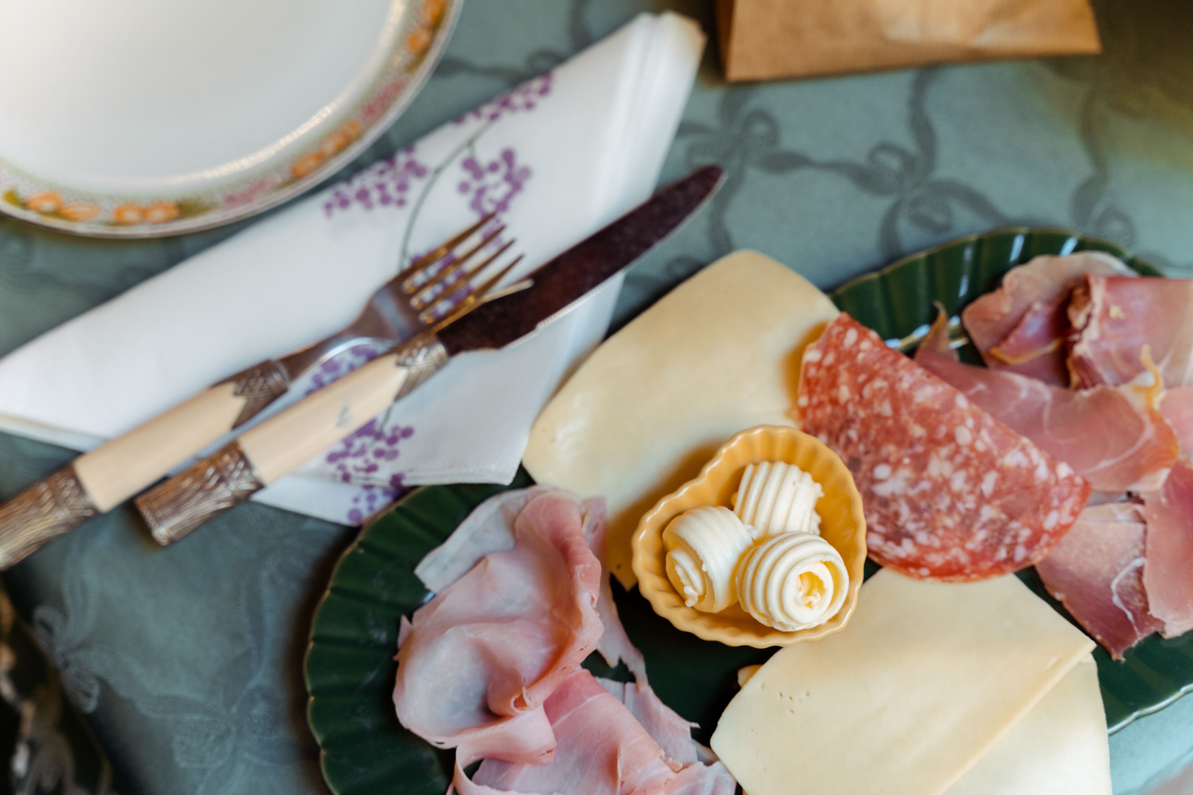 A table with a plate of cheese, cured meats, and a small bowl of cream cheese.