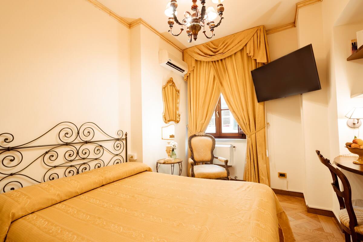 A hotel room with a bed, a chair, a table, a TV, and a window with curtains.