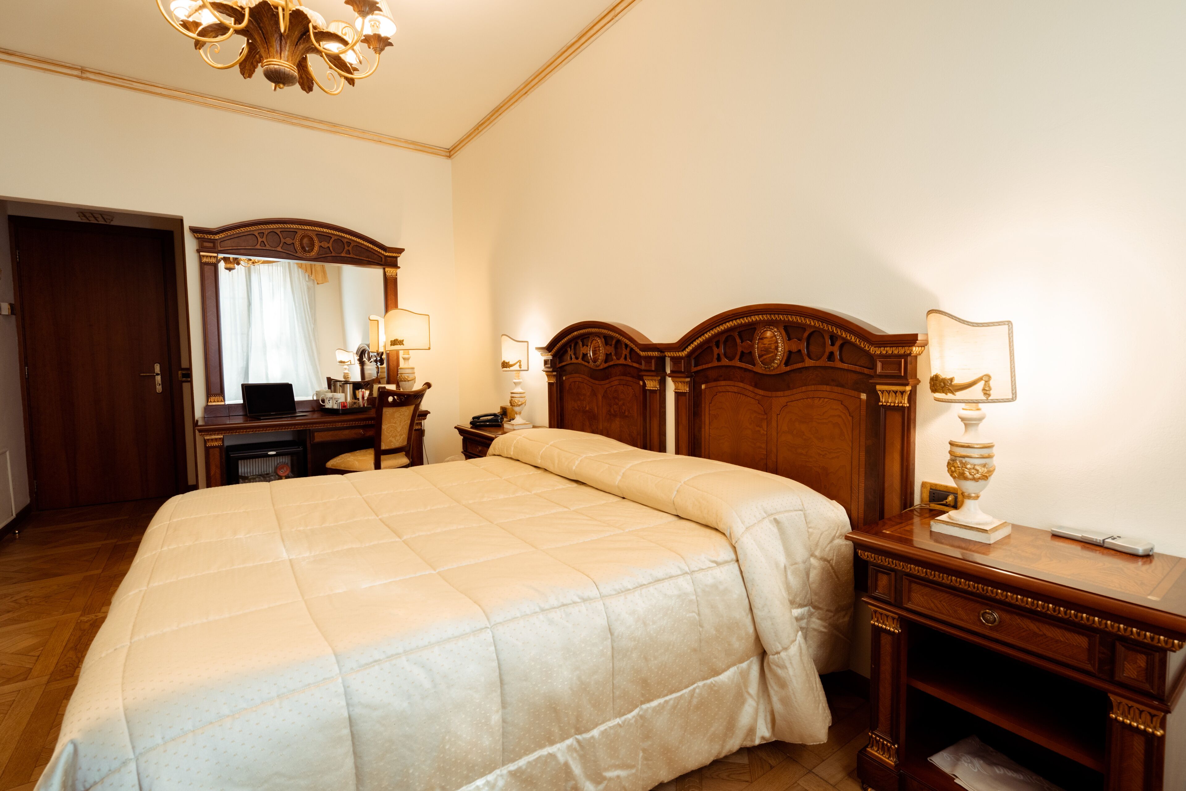 A hotel room with a large bed, wooden headboard, a desk with a lamp, a mirror, and a wardrobe.