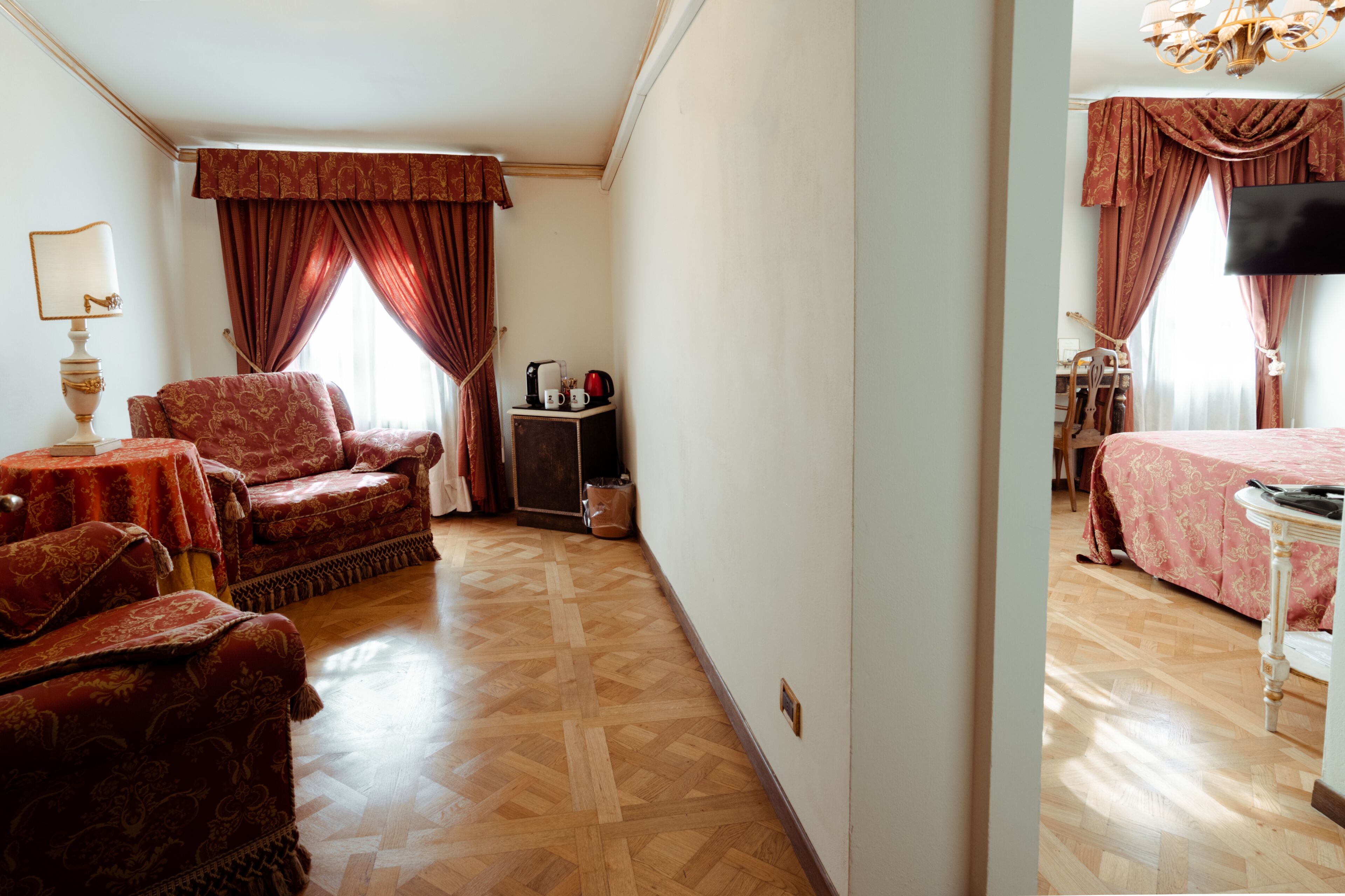Una camera d'albergo con pavimento in parquet, un'area salotto con divano e poltrone, un tavolino con una lampada e un letto con copripiumino rosa.